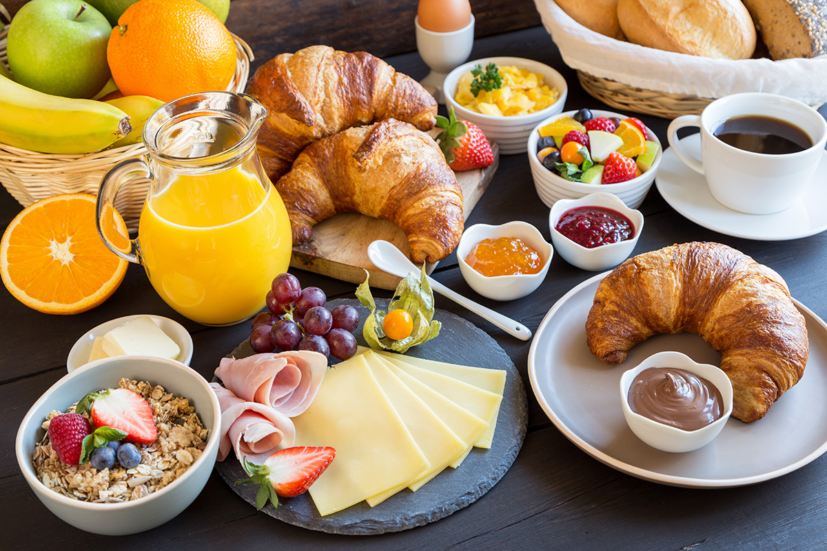A diverse breakfast selection at Hotel Welcome Inn: Croissants, muesli, cheese, ham, fresh fruit, juices, coffee, and egg dishes served on a dark wooden table.